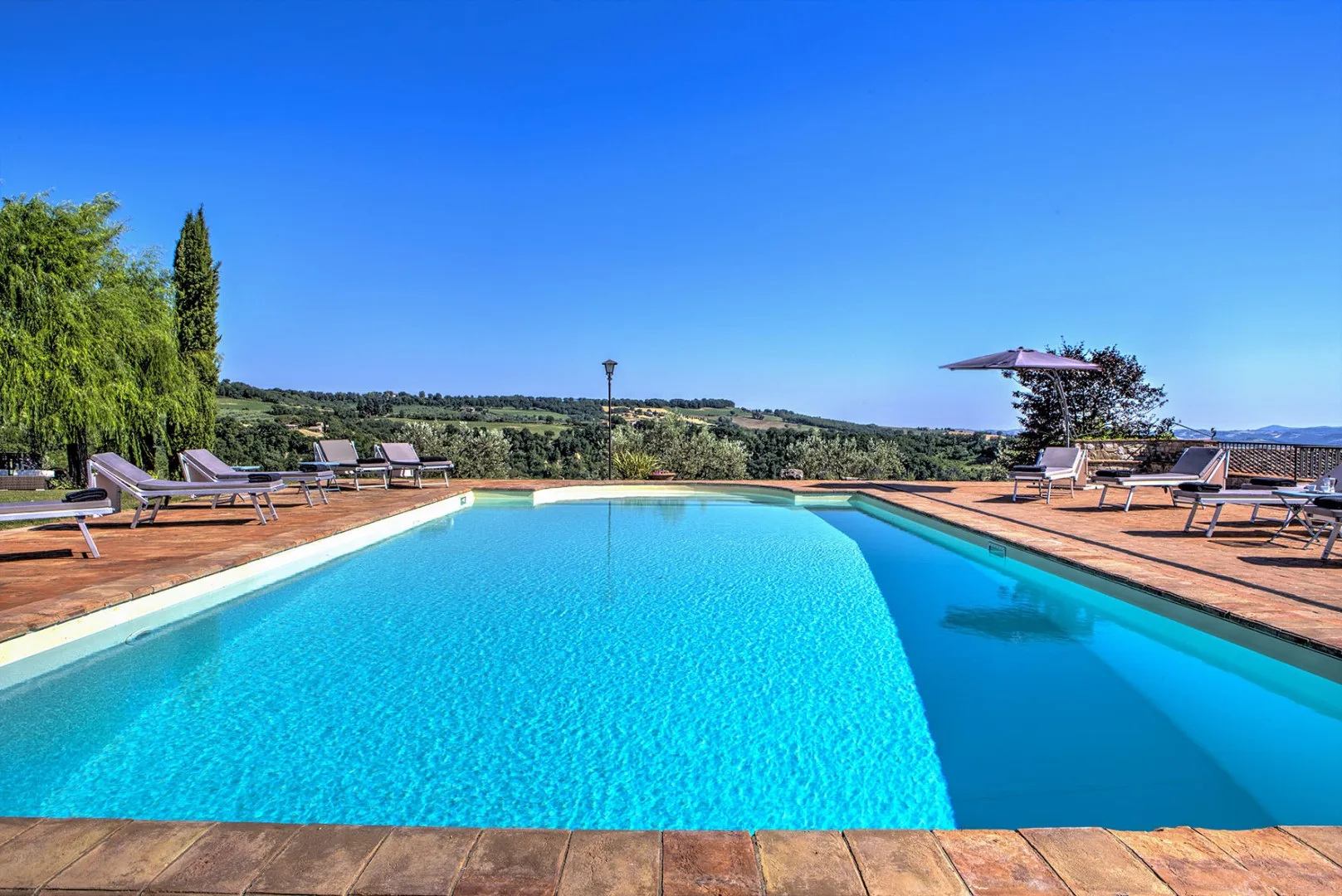 A long outdoor swimming pool is surrounded by a wooden deck, lounge chairs, and a hillside view.
