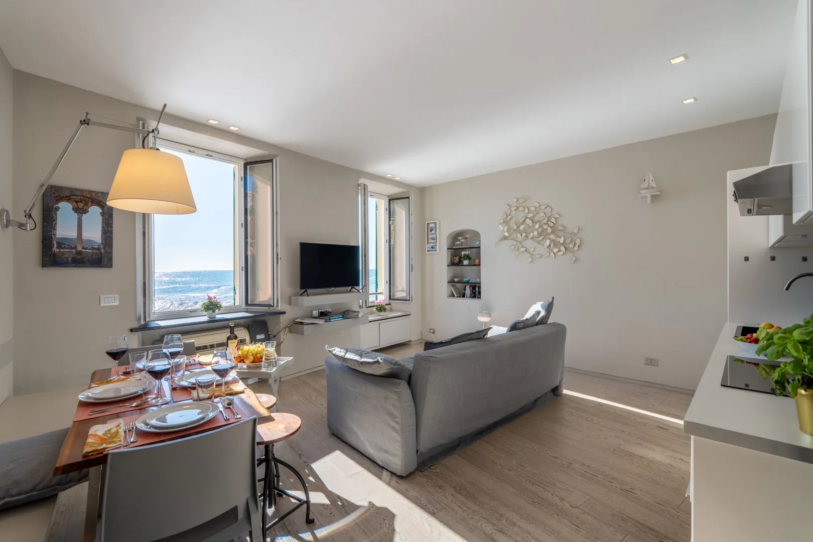 A bright open-plan living and dining room has a gray sofa, set table, and large windows overlooking the sea.