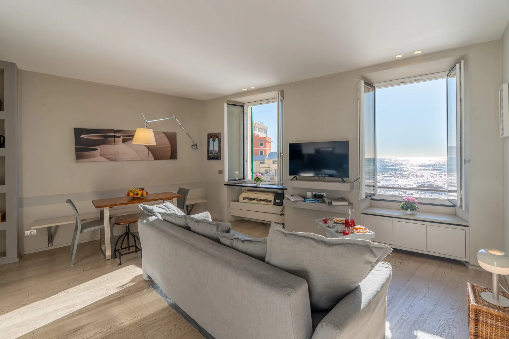 A bright living room has a gray sectional sofa, dining table, and large windows overlooking the sea.