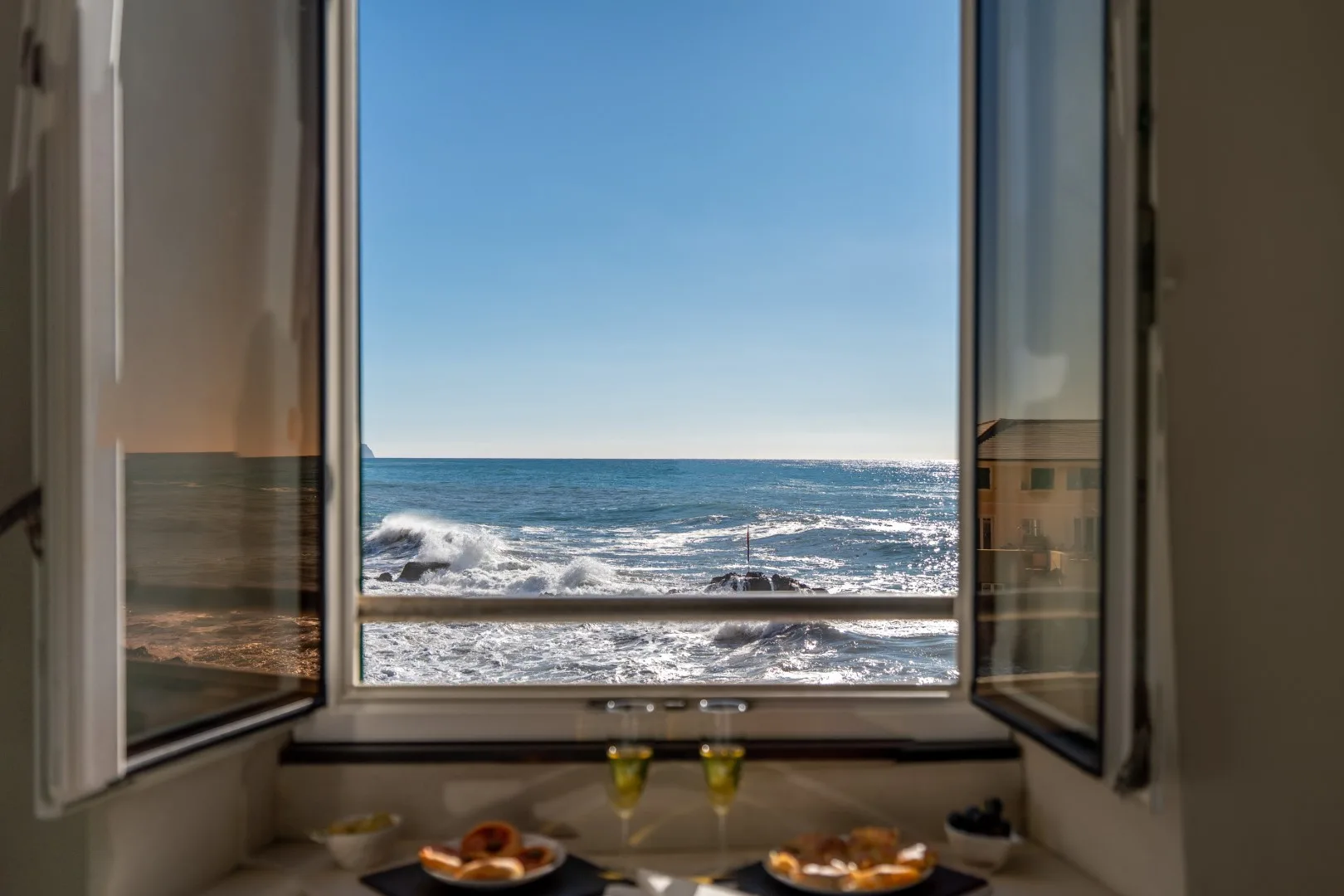 An open window frames ocean waves, with two plates of food and drinks on the sill.