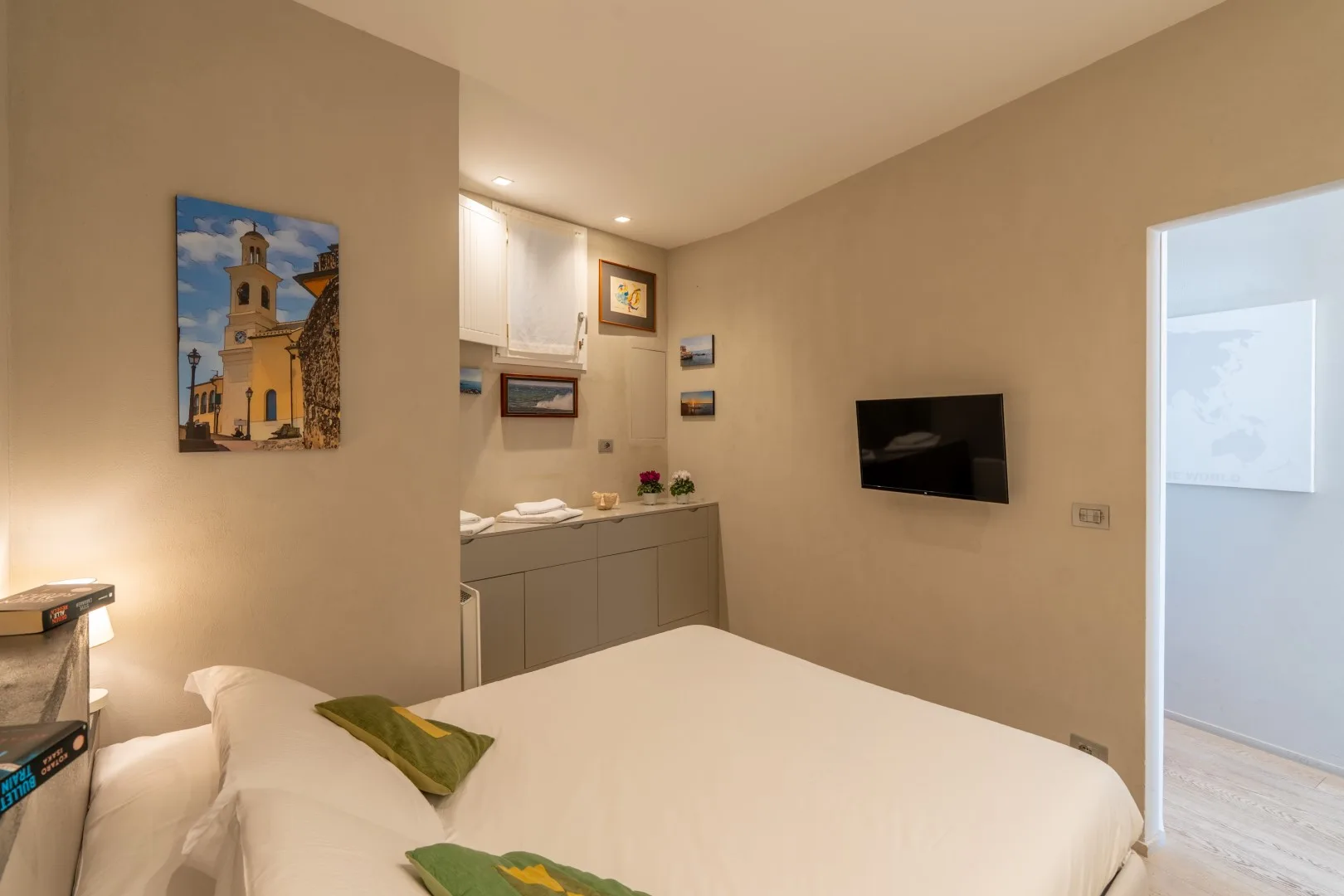 A bedroom with a white bed, green accent pillows, a wall-mounted television, and a small kitchenette.