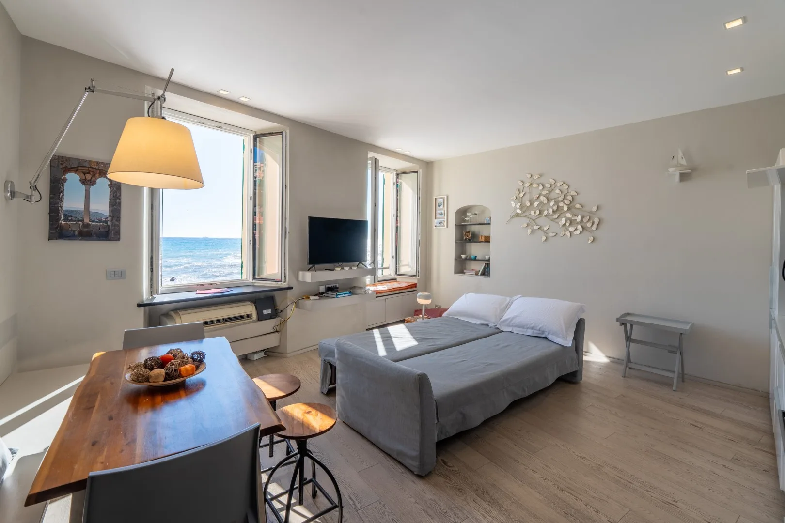 A bright bedroom has a double bed, desk, television, and large windows overlooking the sea.