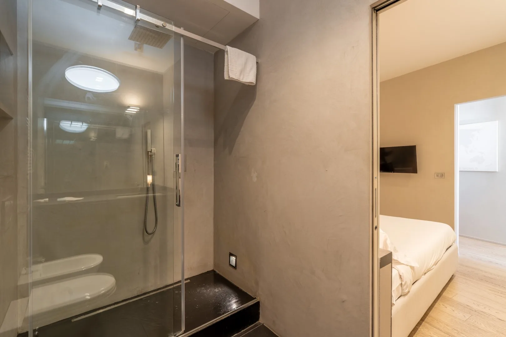 A glass-enclosed shower with a handheld fixture sits beside an open doorway to a bedroom.