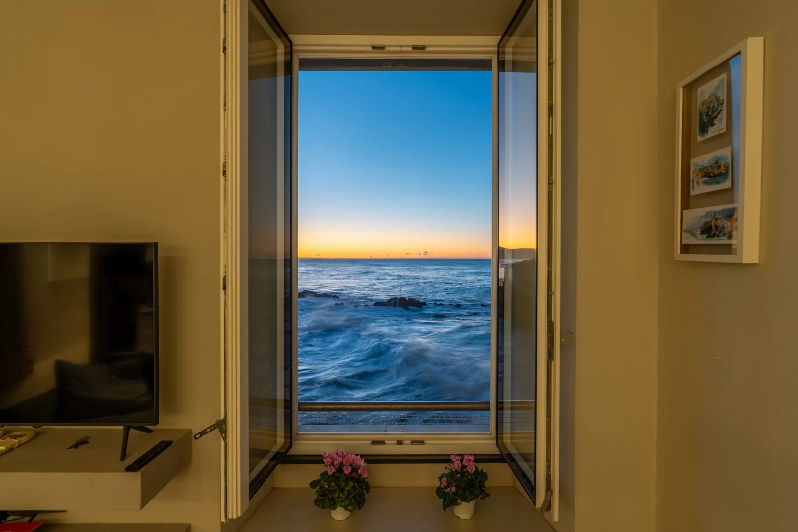 A window opens onto the ocean at sunset, with two potted flowers on the sill and framed pictures on the wall.