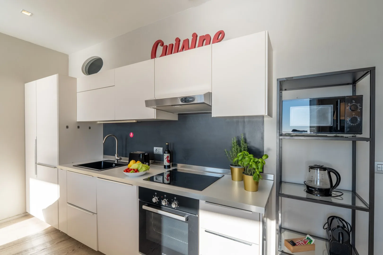 A bright modern kitchen has white cabinets, a black backsplash, a cooktop, and countertop plants.