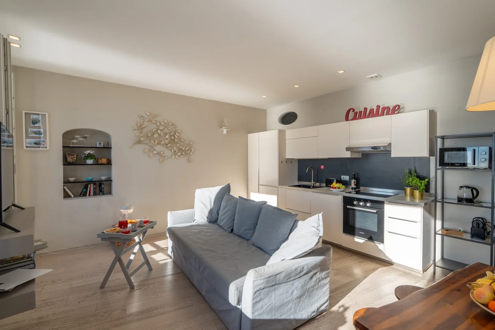 A bright open-plan living room and kitchen features a gray sofa, small coffee table, and white cabinets.