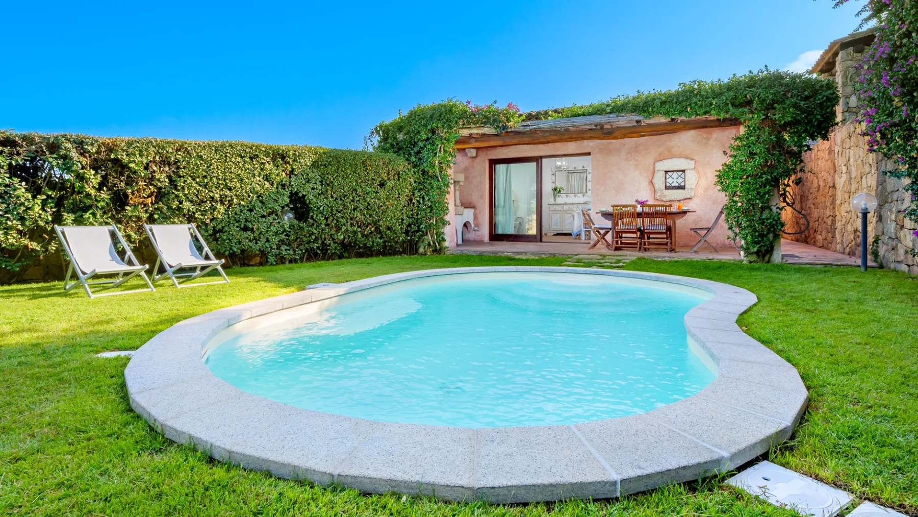 Una piscina en el jardín junto a una pequeña construcción, rodeada de césped y setos.
