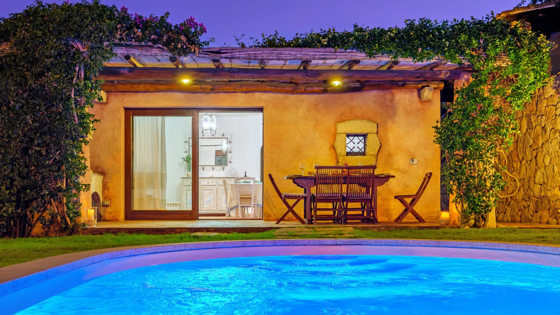 Una piscina exterior iluminada está junto a un edificio pequeño con puertas de vidrio.