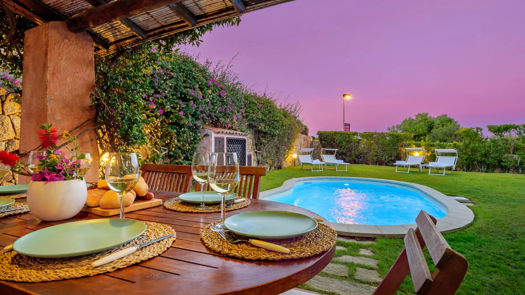 Una mesa de comedor al aire libre da a una piscina iluminada y un jardín al anochecer.