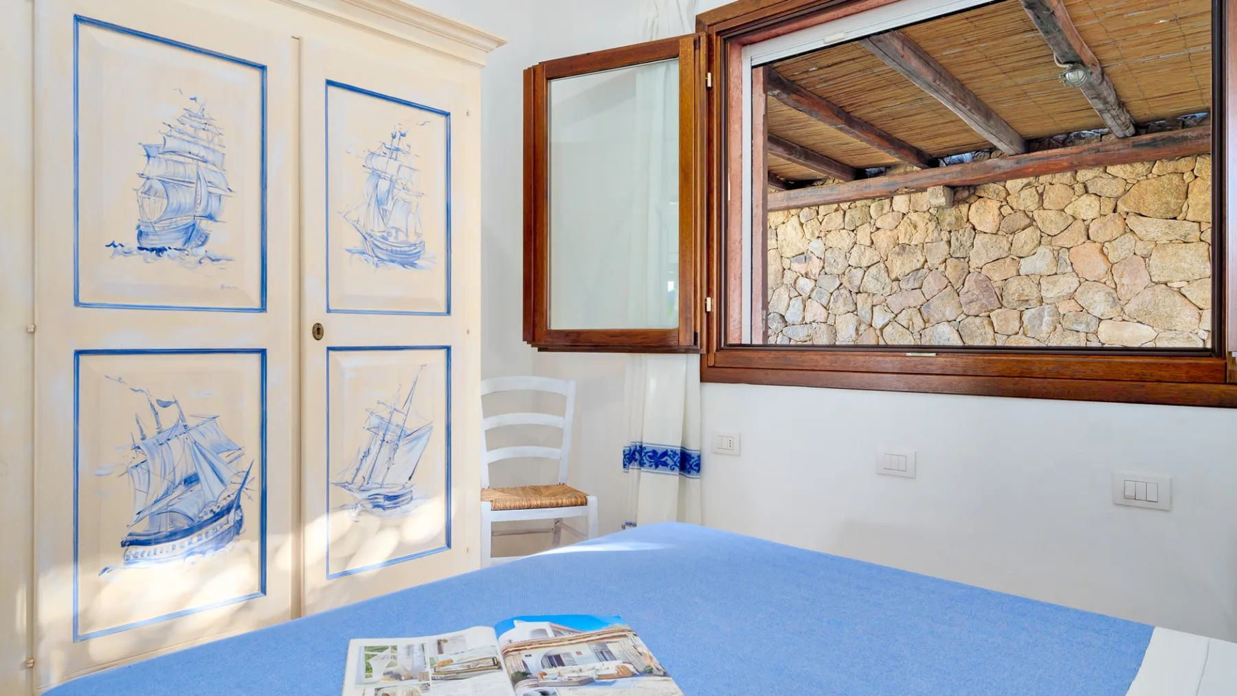 Una habitación luminosa con una cama cubierta de azul, láminas de barcos y una ventana frente a un muro de piedra.