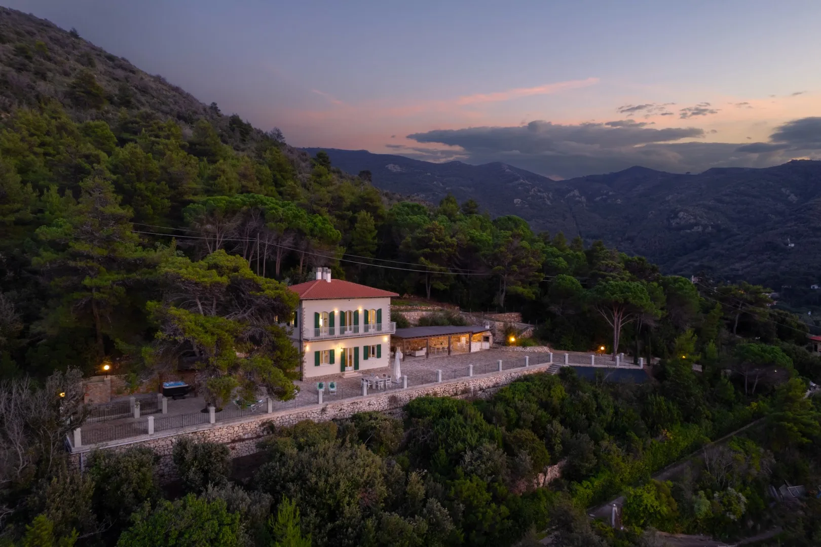A building sits on a wooded hillside overlooking a mountain valley at dusk.