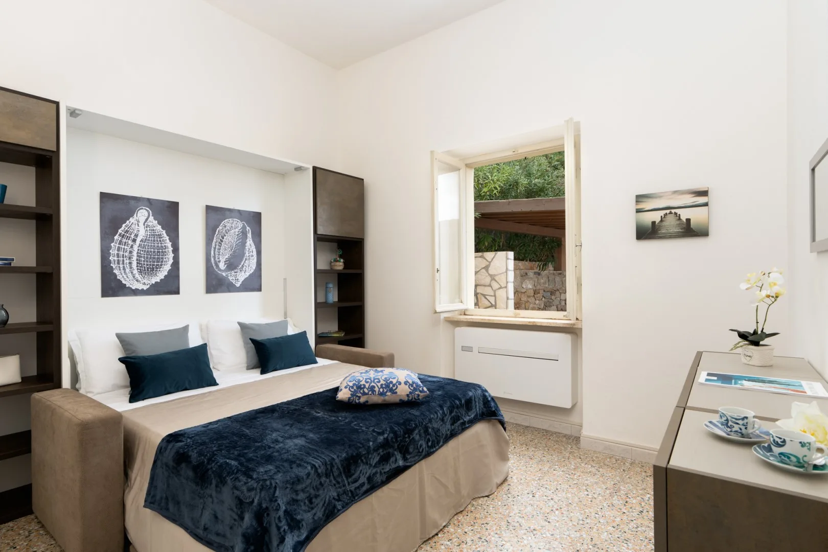 A bright bedroom with a bed beneath two framed shell prints and a window.