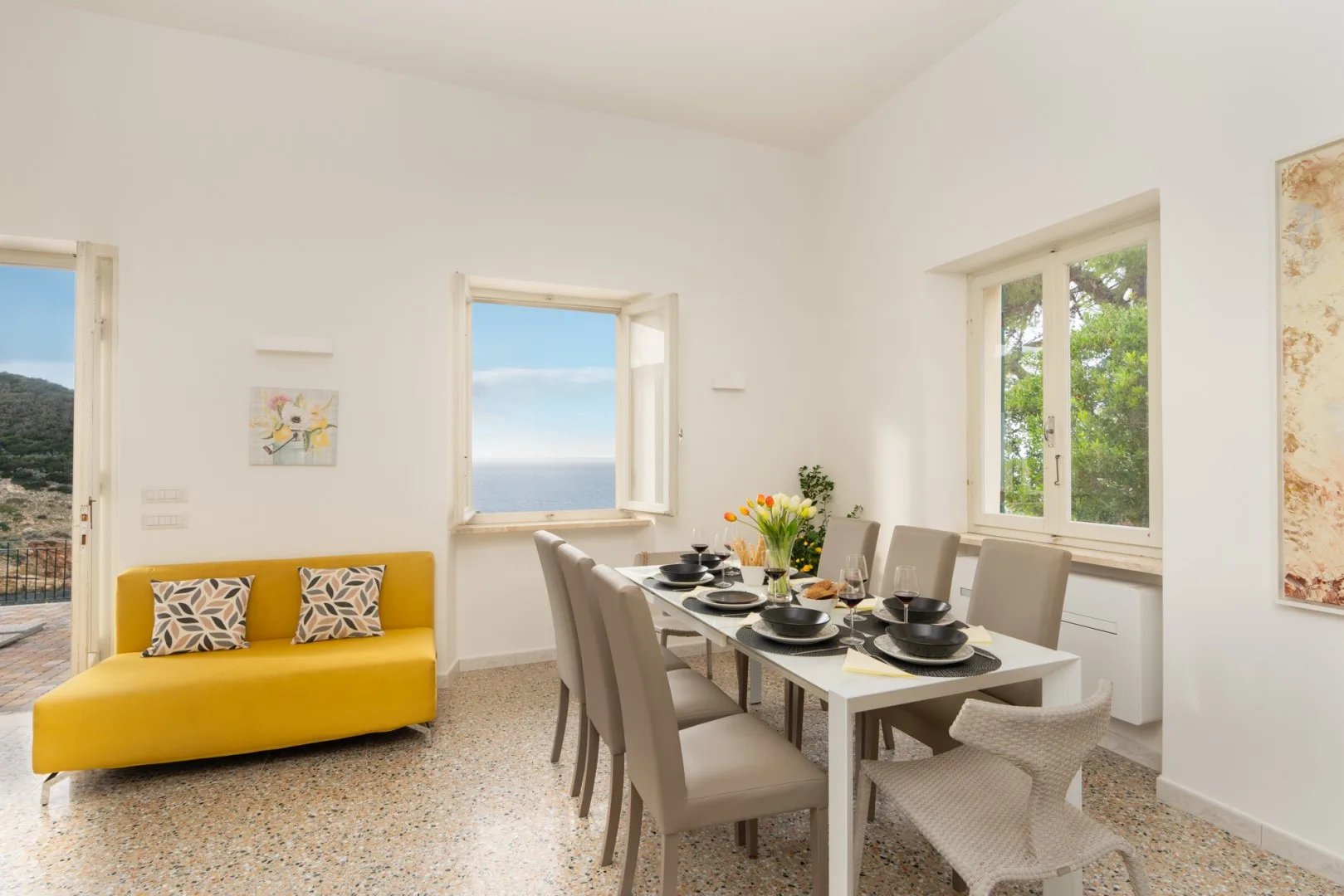A bright dining area with a set table beside a yellow sofa and open windows.