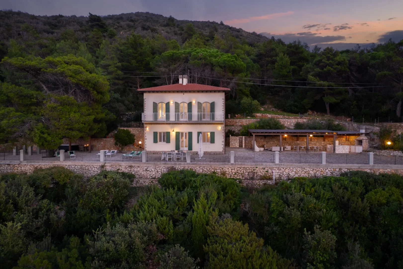 A house and adjoining patio sit among dense greenery beneath wooded hills at dusk.