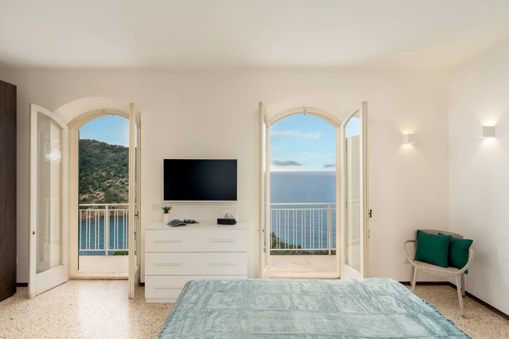 A bedroom with a bed, a television, and open doors overlooking the sea.