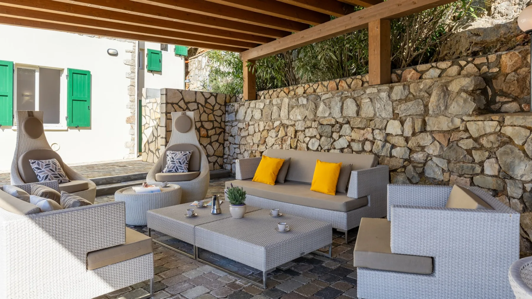 An outdoor seating area beneath a wooden pergola, with cushions beside a stone wall.