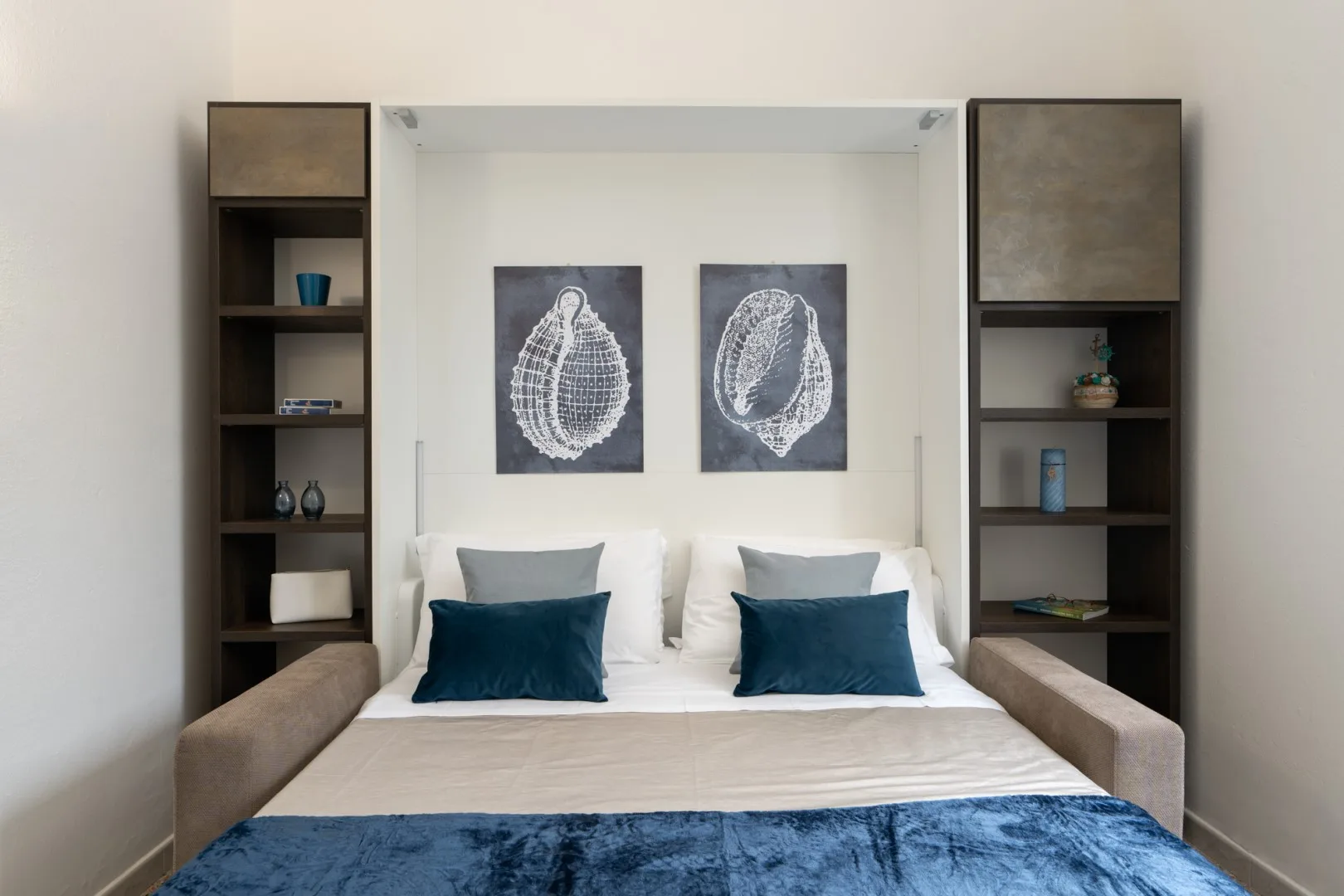 A neatly made bed sits between two dark shelving units, with framed prints above.
