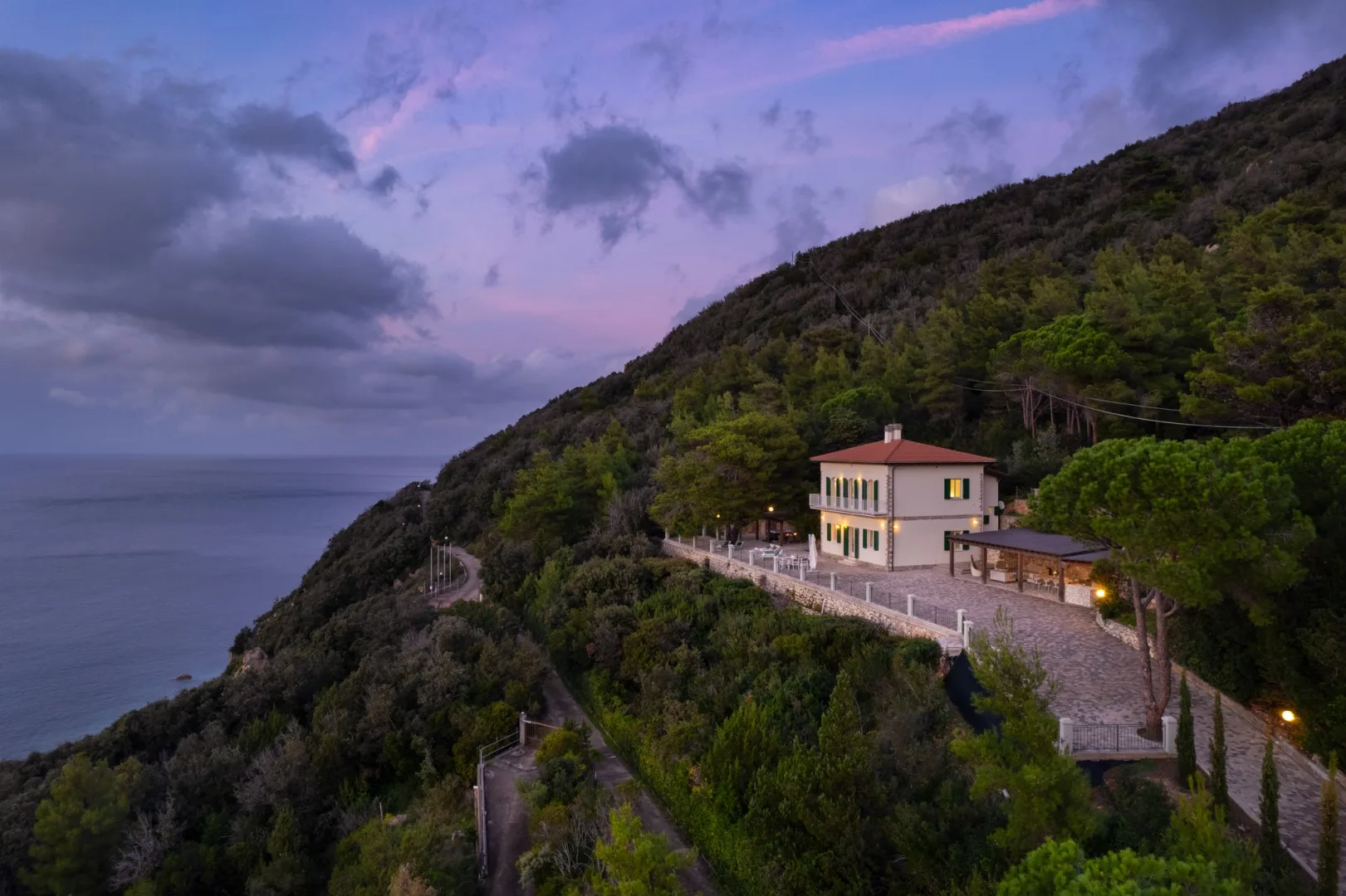 A hillside house overlooks the sea at dusk, with a lit terrace.