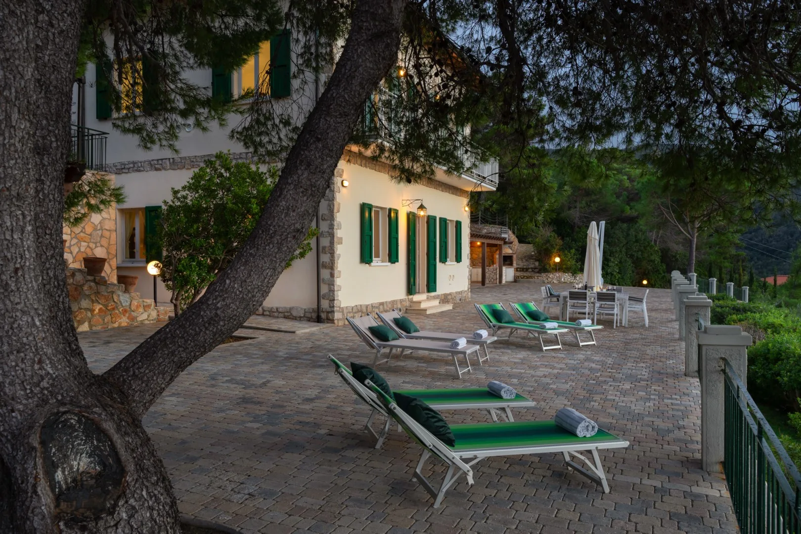A stone-paved outdoor terrace beside a white building, with green lounge chairs and trees.
