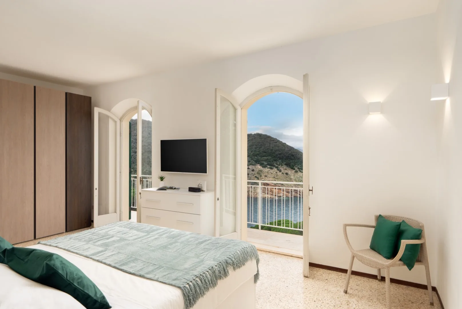 A bedroom with a bed, a dresser, and open doors overlooking a green hillside.