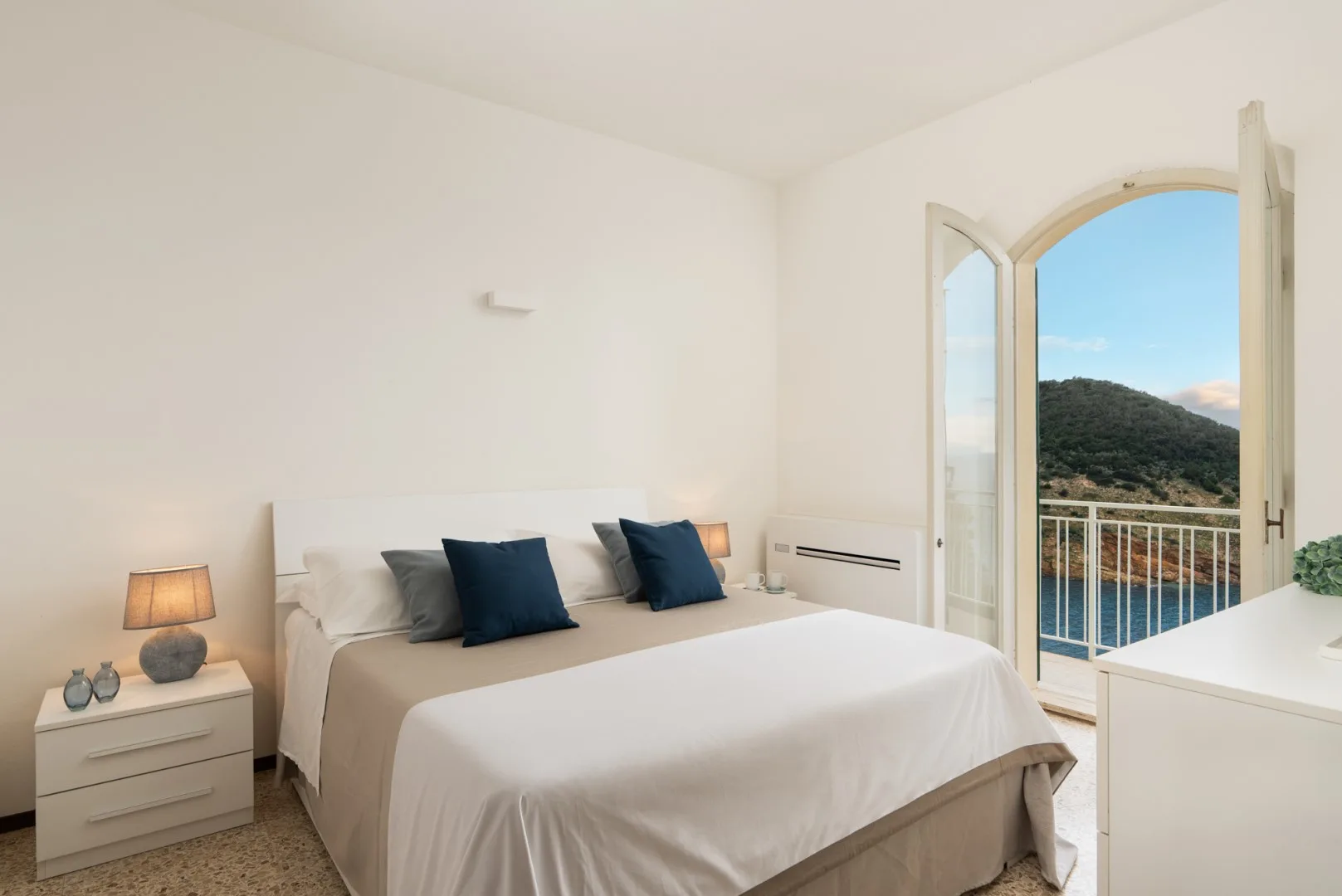 A bright bedroom with a neatly made bed beside open balcony doors.