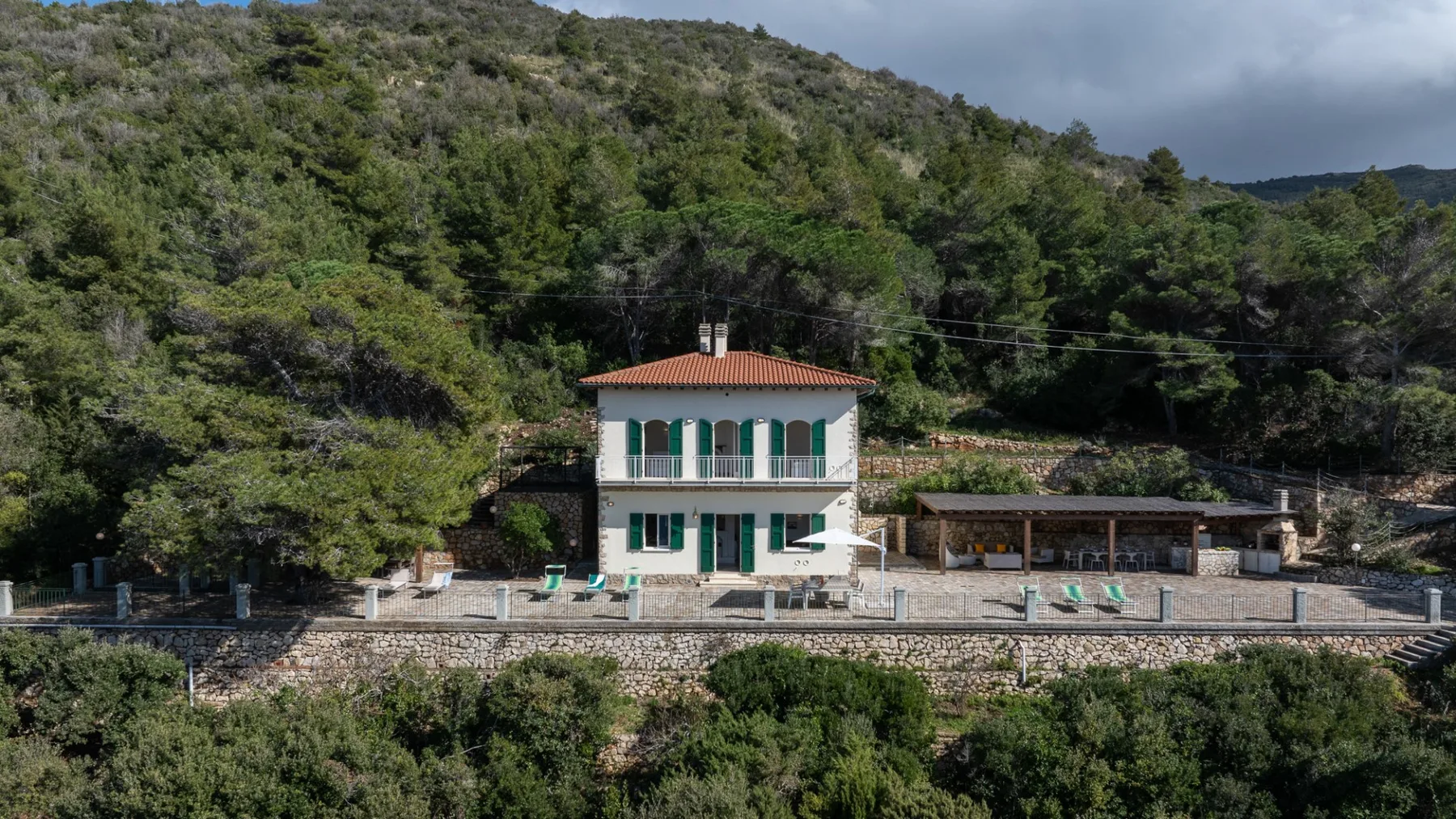 A house with green shutters stands among wooded hills above a stone retaining wall.