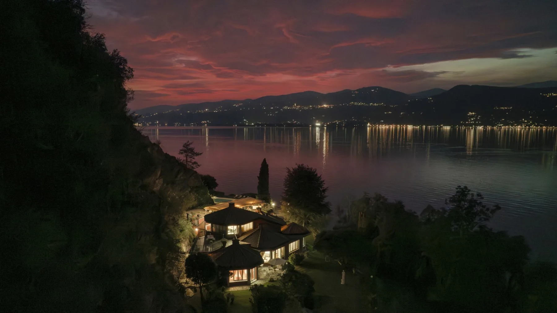 A lakeside house glows beneath a red sunset, with distant lights reflected on the water.