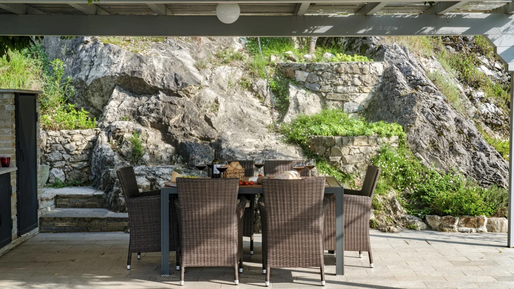 An outdoor dining area with woven seating sits beside a rocky, planted hillside.