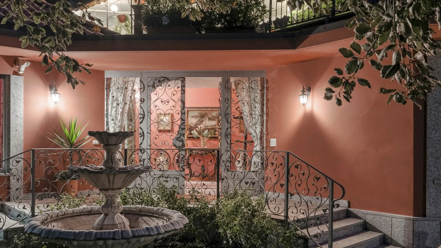 A warmly lit courtyard features a decorative fountain, plants, and a wrought-iron railing.