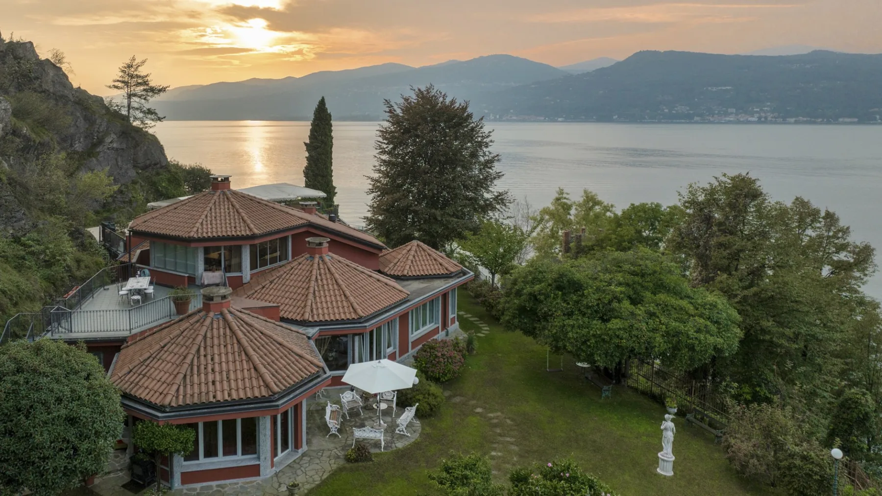 A hillside house overlooks a lake and surrounding mountains at sunset.