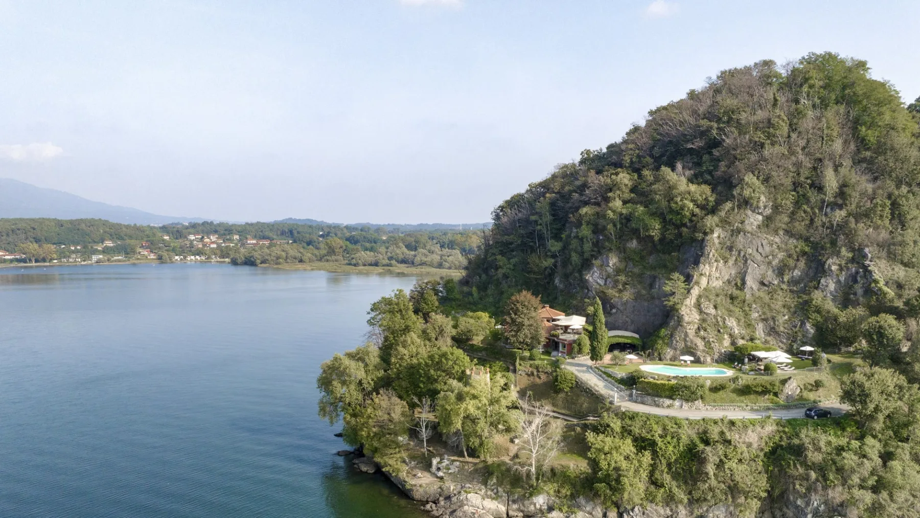 A lake curves around a wooded rocky shoreline beneath a green hillside.