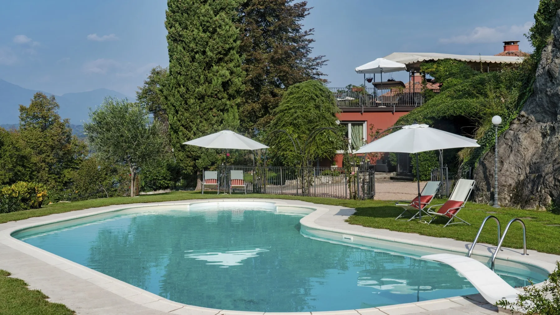 A swimming pool surrounded by greenery, with umbrellas and lounge seating nearby.