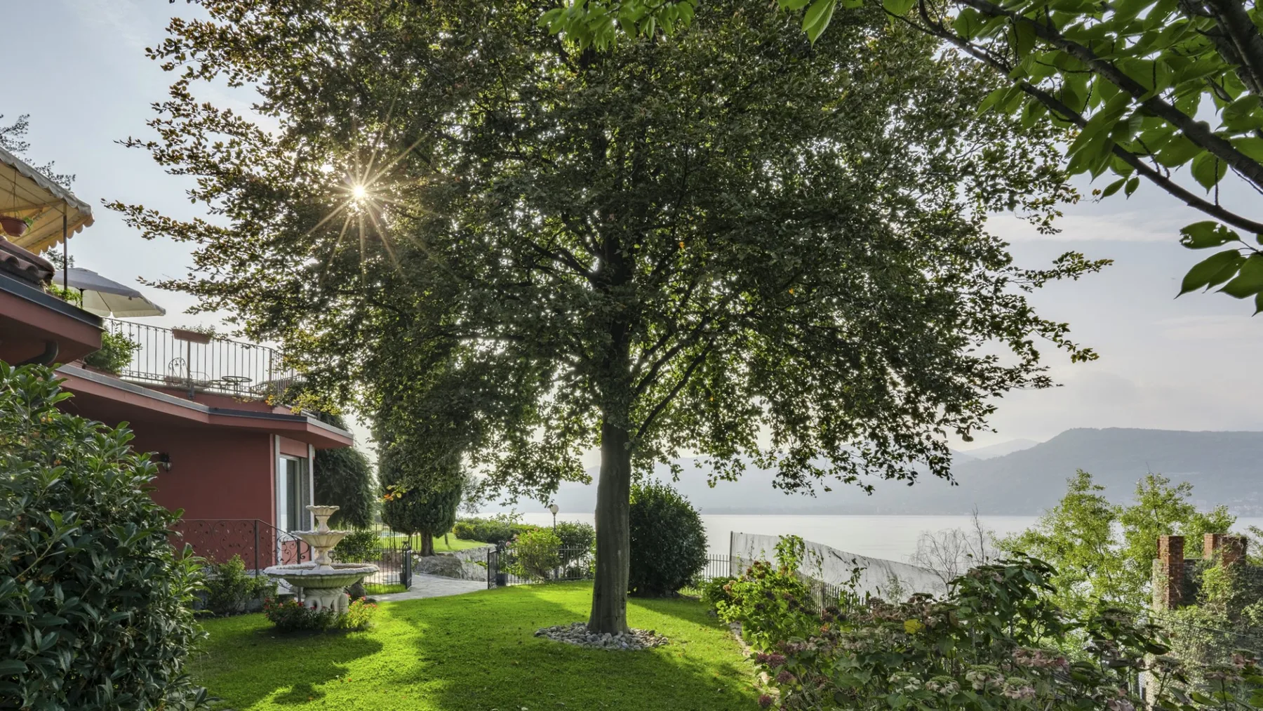 A leafy garden beside a red house overlooks a lake and distant hills.