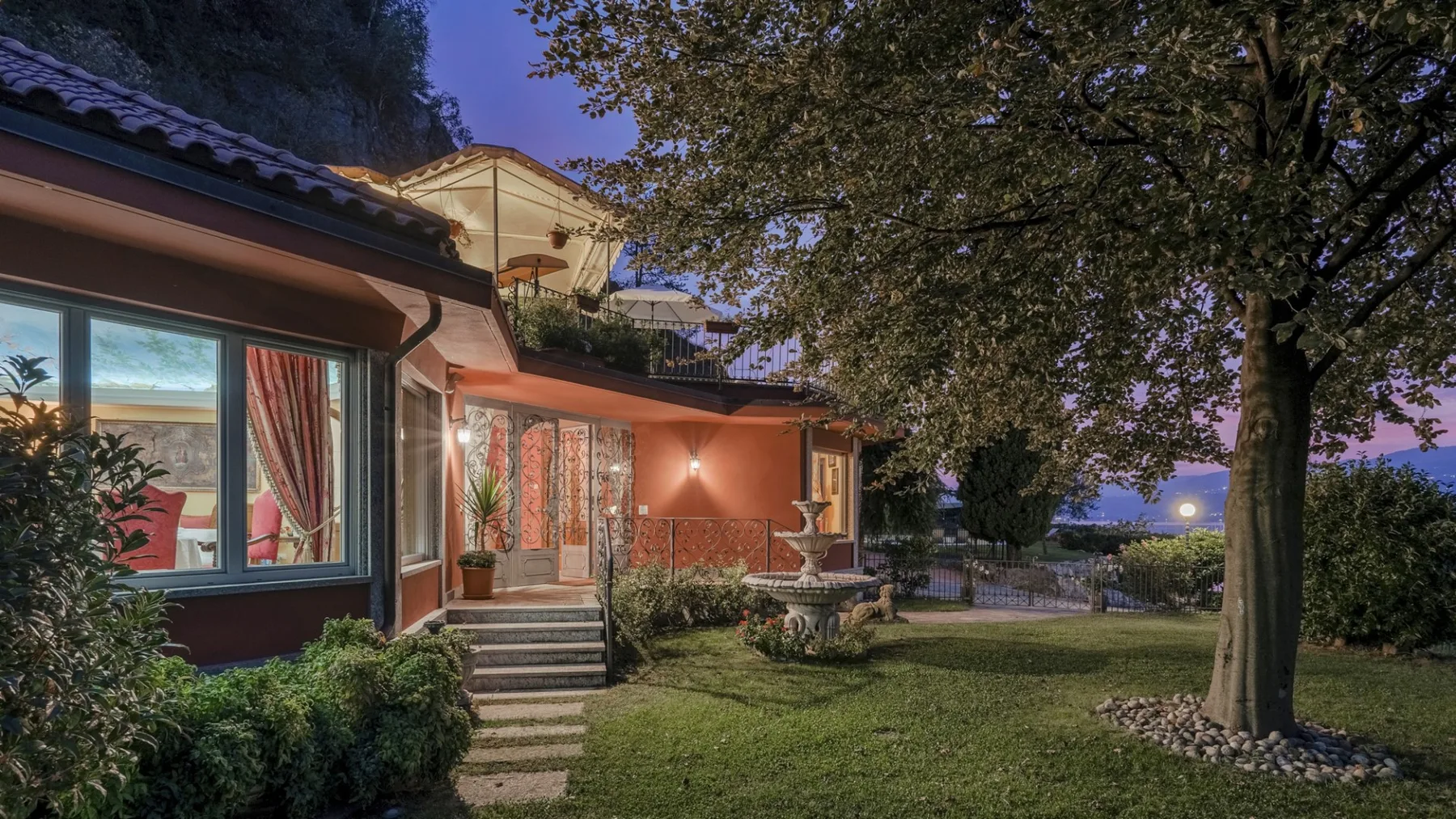 A house and garden at dusk, with a tree and outdoor fountain.