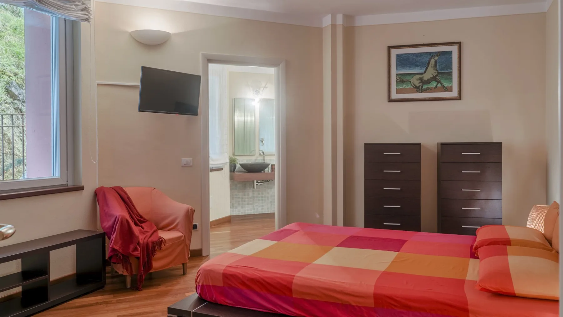 A bedroom with a colorful bedspread, a chair, and two dressers.