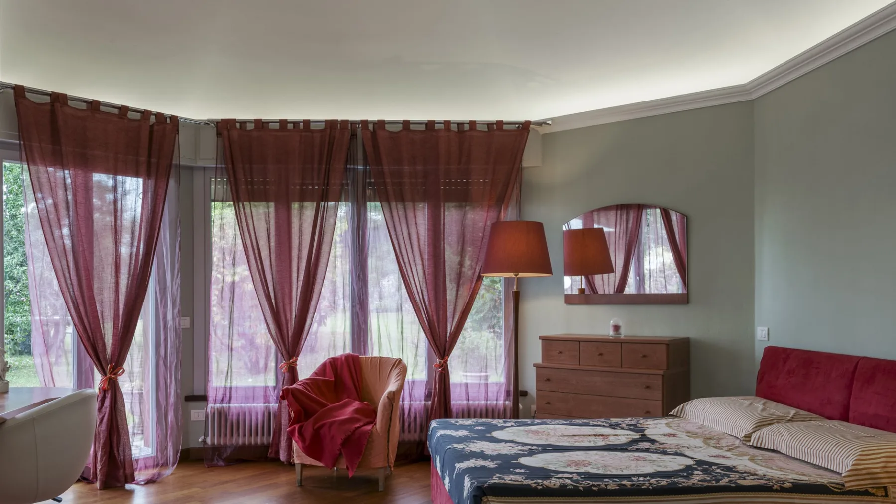 A bedroom with a bed, a dresser, and tall windows with sheer curtains.