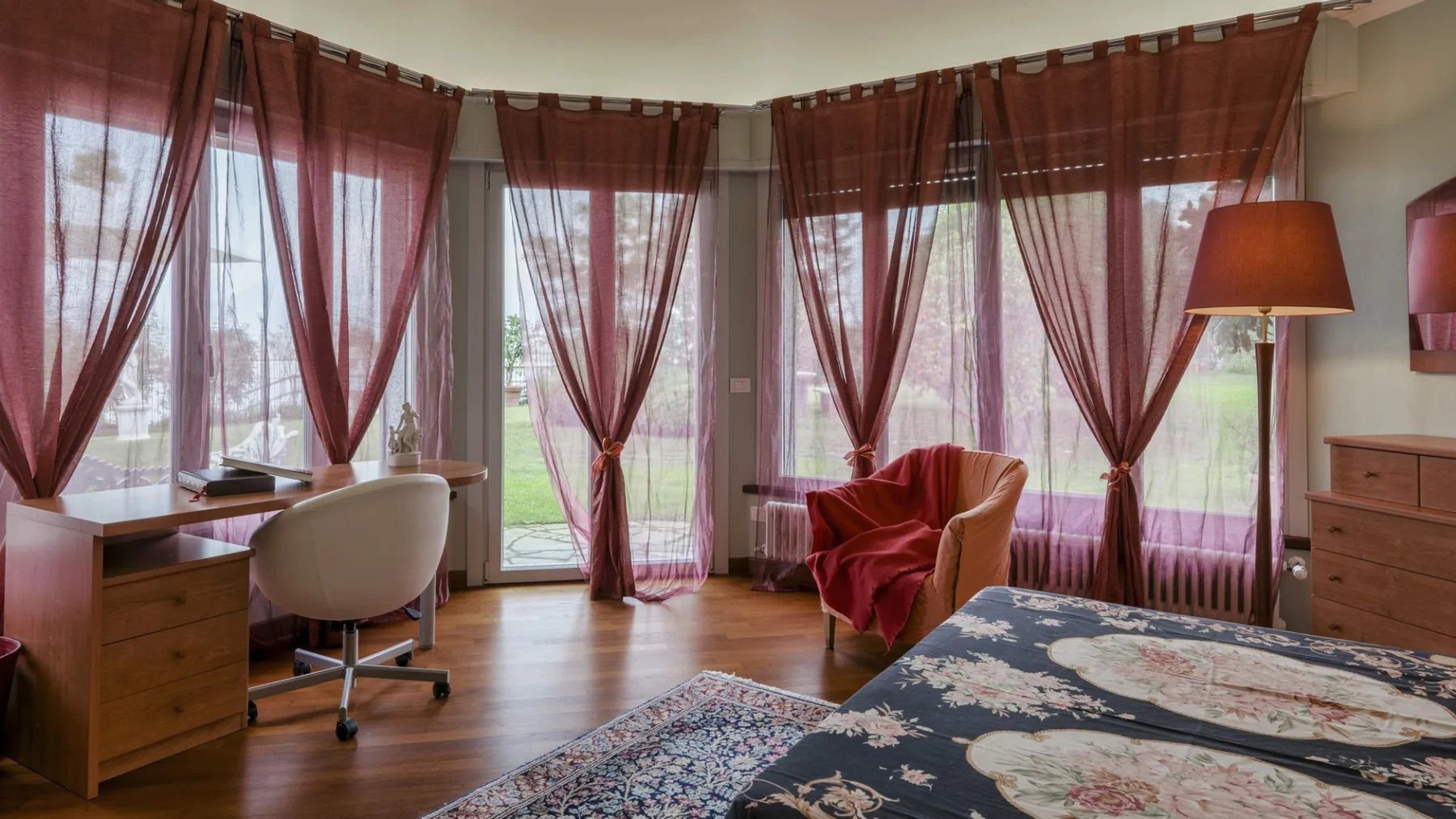 A bedroom with a desk, seating, and tall windows draped in sheer curtains.