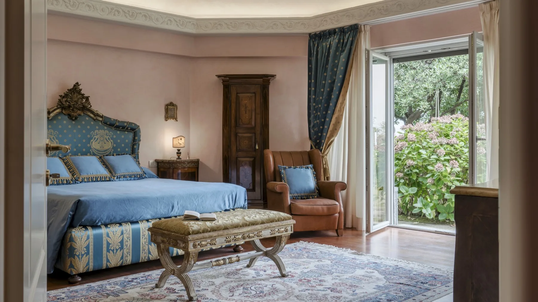 A bedroom with a blue bed, an armchair, and an open glass door to the garden.