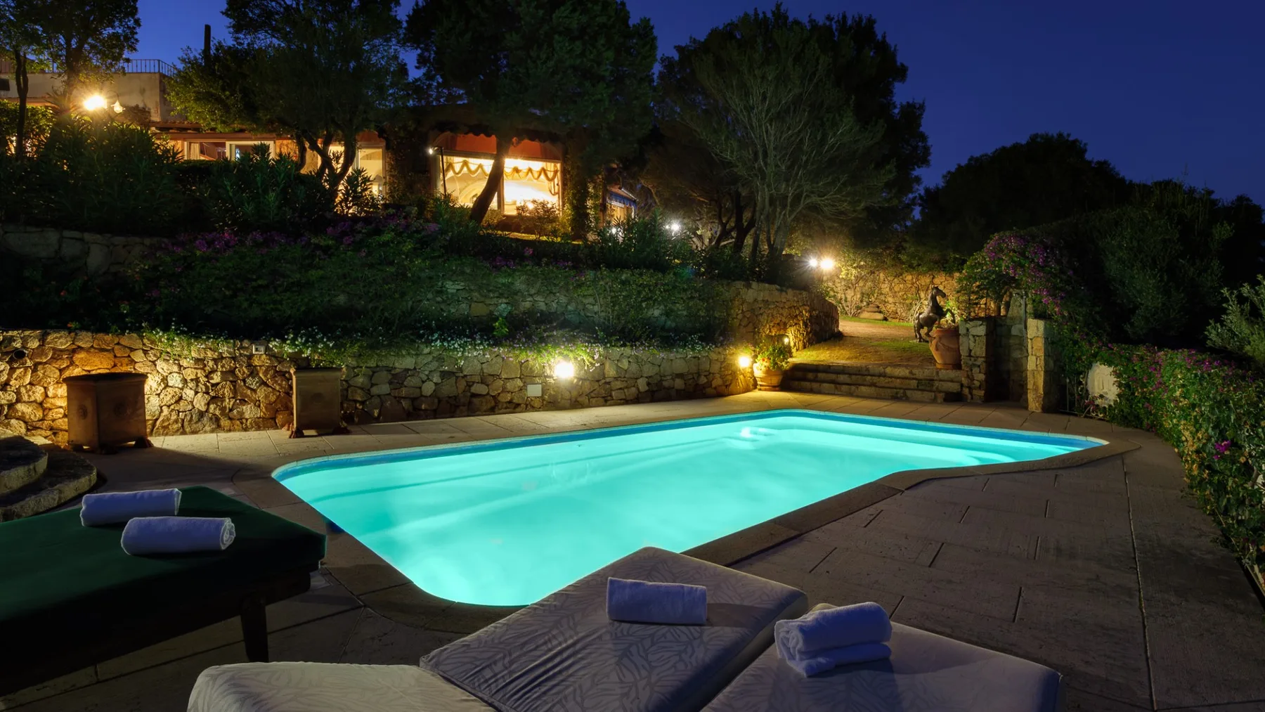 Una piscina exterior iluminada se encuentra entre senderos de piedra y vegetación por la noche.