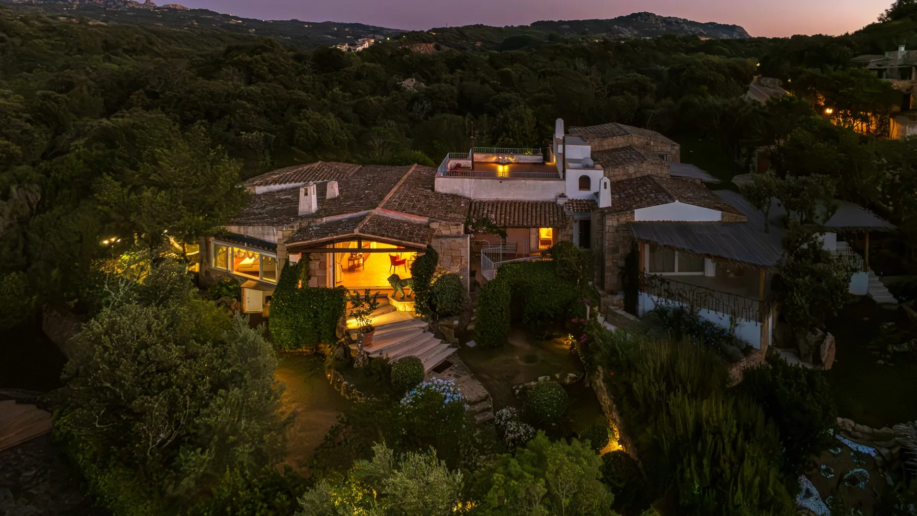 Una casa iluminada cálidamente está entre árboles densos en una ladera al anochecer.