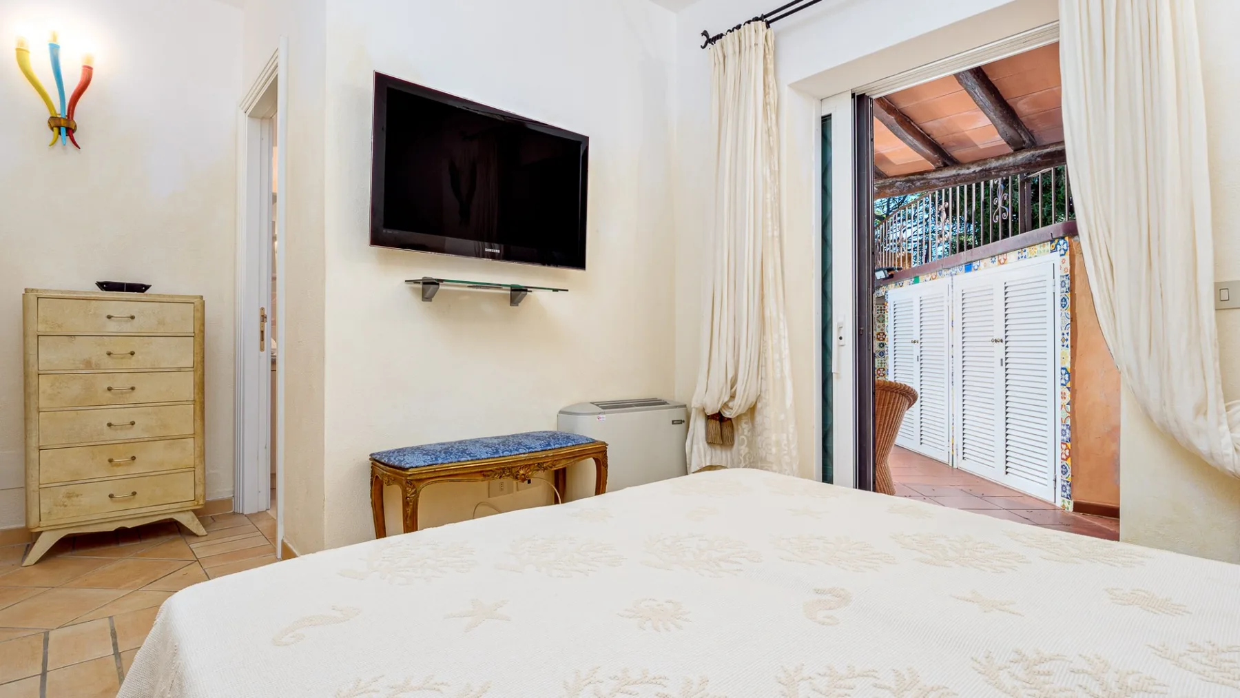 Un dormitorio luminoso con cama, televisor y una puerta abierta a un balcón.
