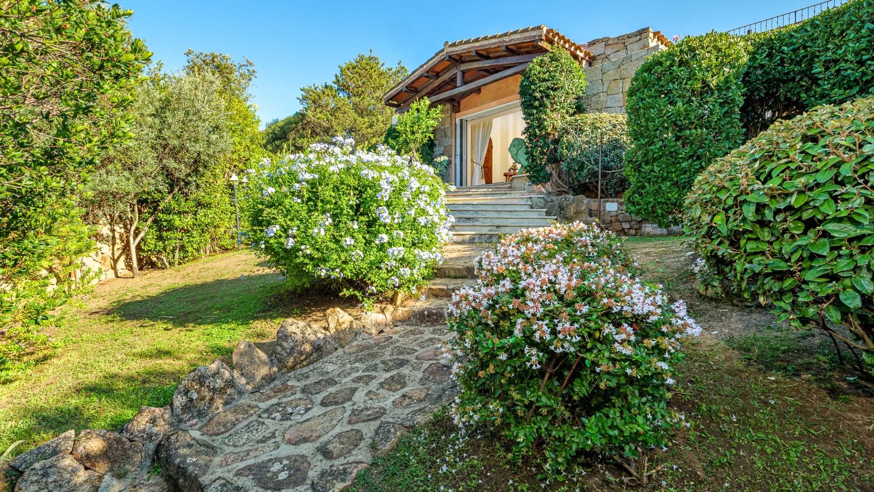 Un sendero del jardín conduce a la entrada de una casa de piedra, rodeada de arbustos floridos y vegetación.