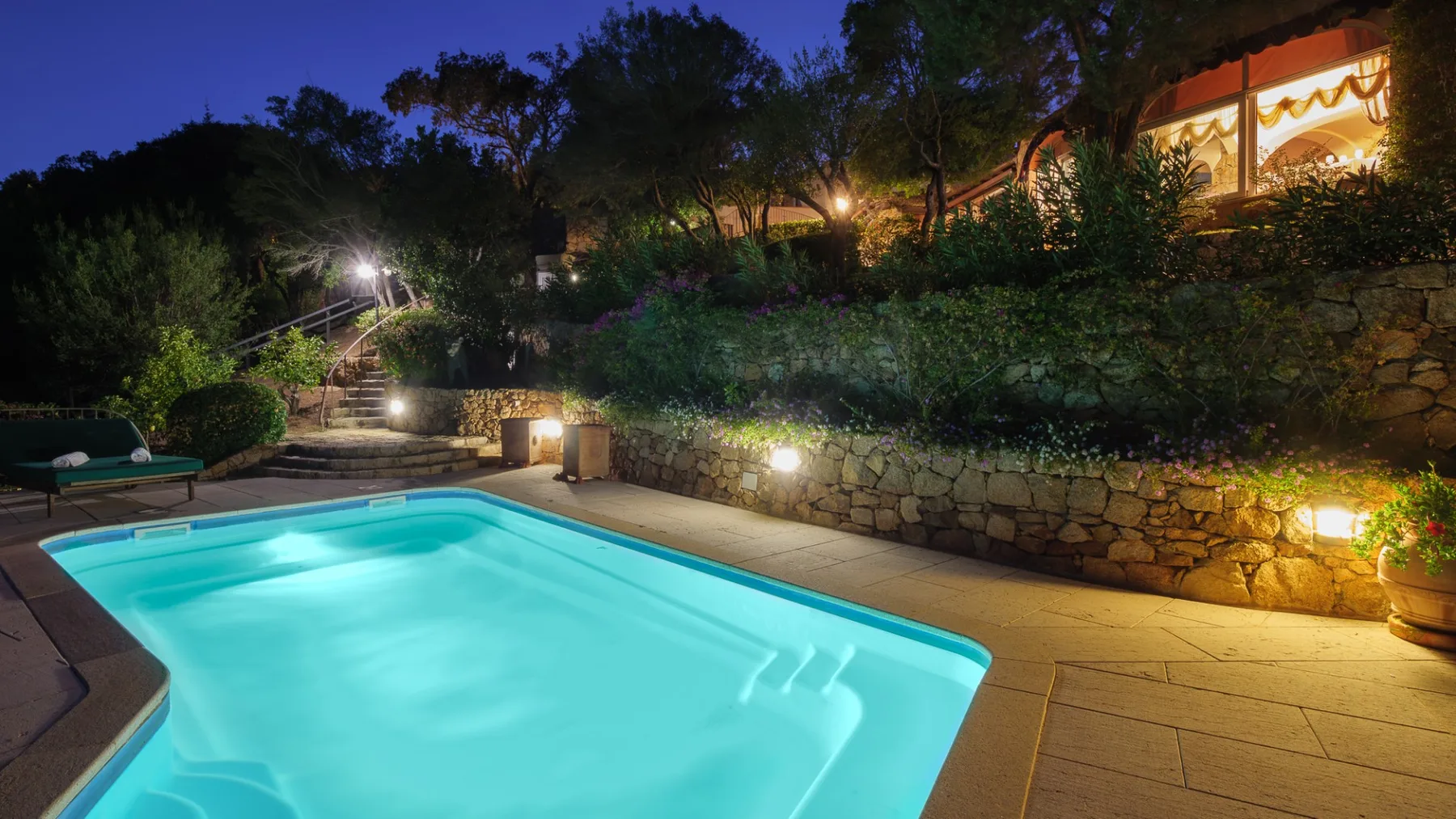 Una piscina exterior iluminada está junto a un muro de piedra en un jardín nocturno.
