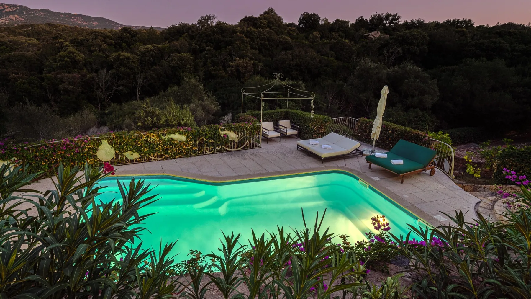 Una piscina al aire libre está rodeada de un jardín, con colinas boscosas al fondo.