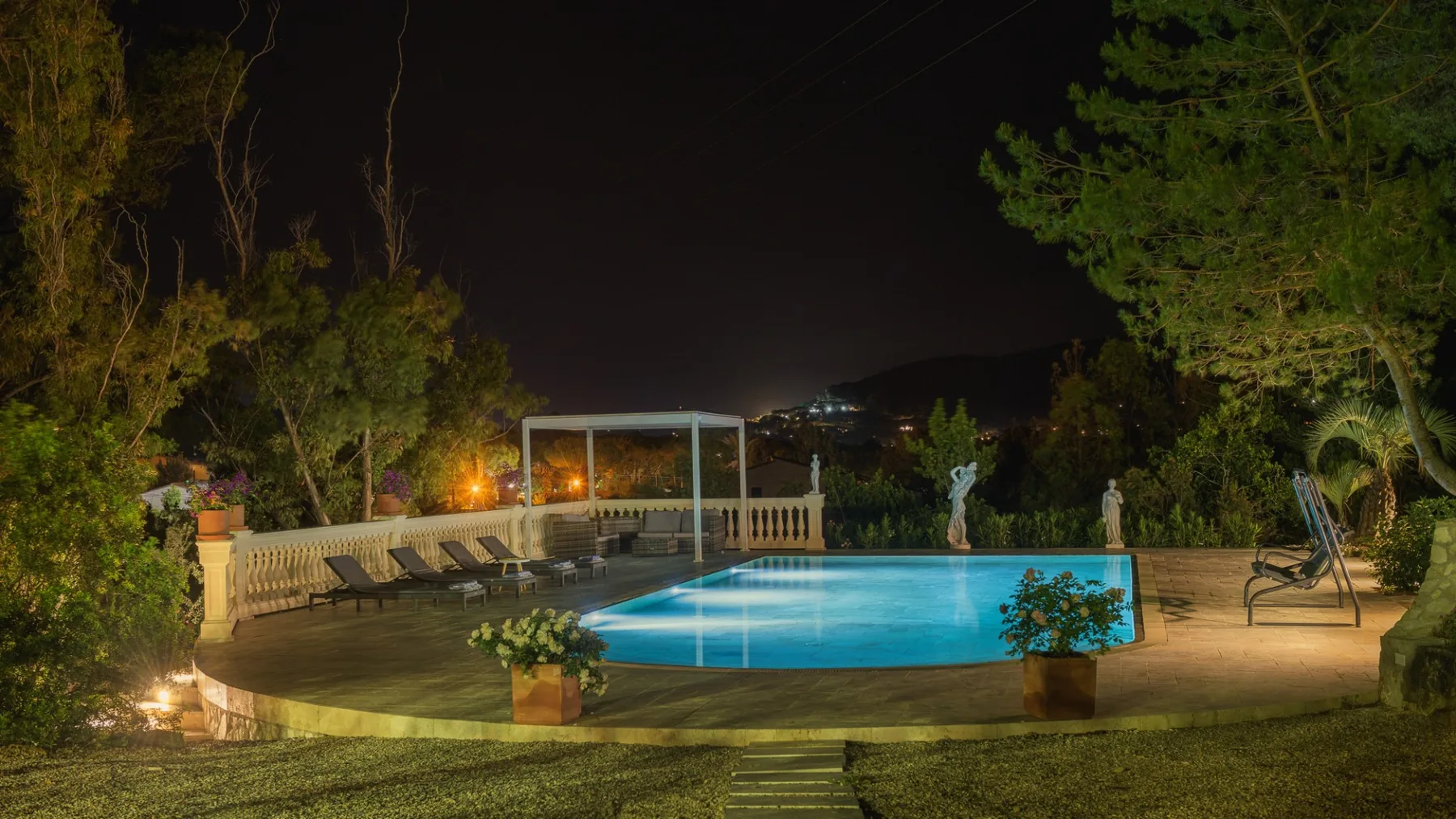 Una piscina iluminada por la noche junto a un patio pavimentado y árboles.