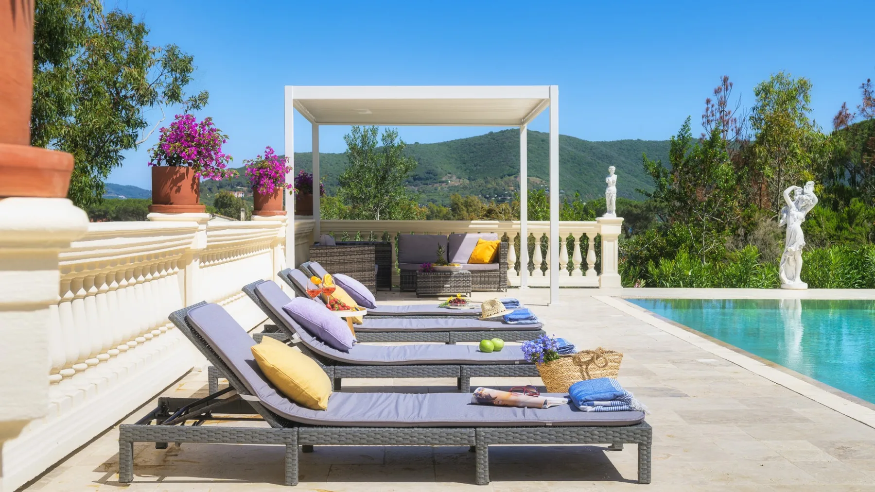 Una terraza soleada junto a la piscina tiene asientos reclinables bajo una pérgola blanca.