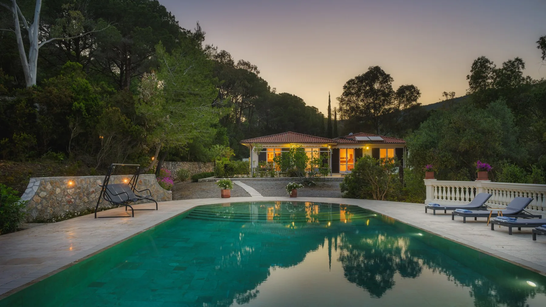 Una piscina refleja los árboles del entorno junto a un patio iluminado al anochecer.