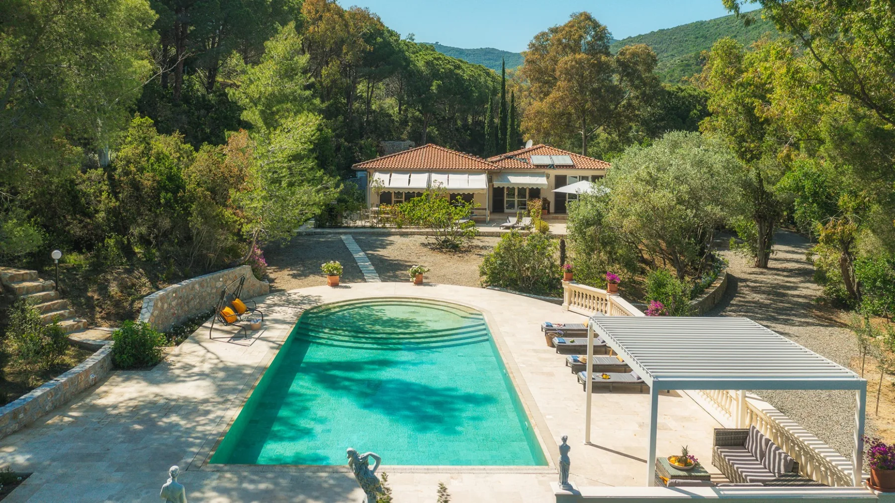 Una piscina y un patio pavimentado están frente a una casa entre colinas boscosas.