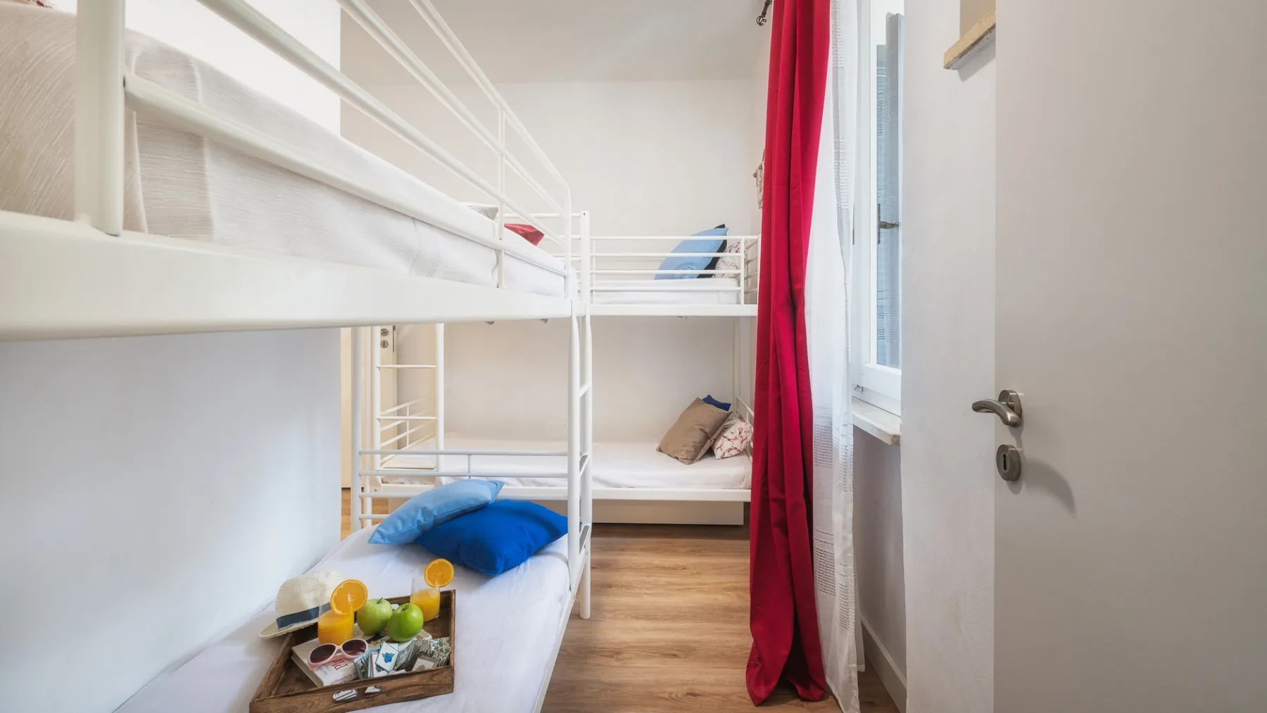 Un dormitorio luminoso con literas blancas, ropa de cama colorida y una cortina roja.
