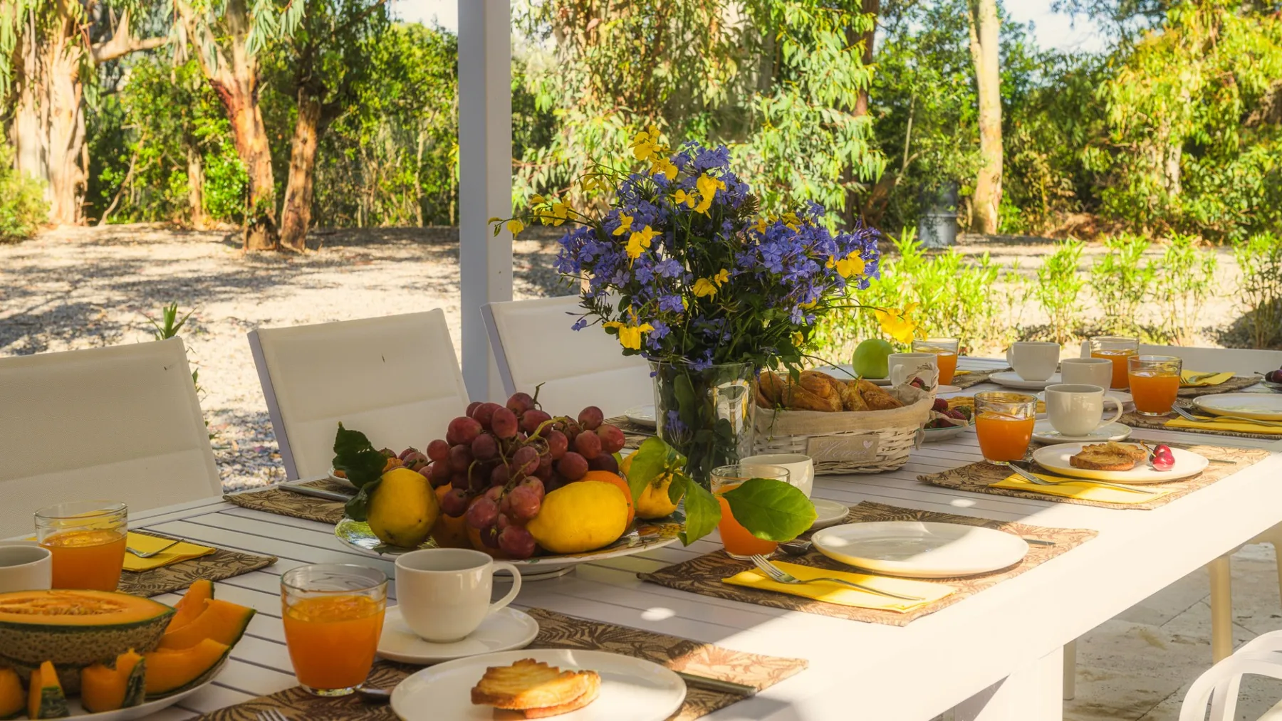 Una mesa de desayuno está preparada al aire libre con fruta, bebidas y flores.